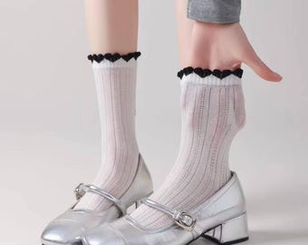 May include: White ribbed socks with a black heart-shaped trim, paired with silver Mary Jane shoes. The shoes have a block heel and a buckle closure. The socks are worn on the legs.