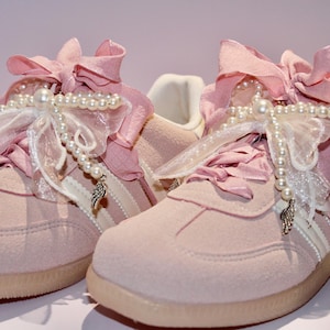 May include: A pair of pink suede sneakers with white stripes and pearl embellishments. The sneakers have pink ribbon laces and white lace accents.