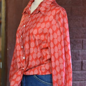 May include: A red shirt with a pattern of light pink teardrop shapes. The long-sleeved shirt is knotted at the waist and paired with blue denim jeans. White buttons fasten the front and cuffs. The shirt is a vintage style.