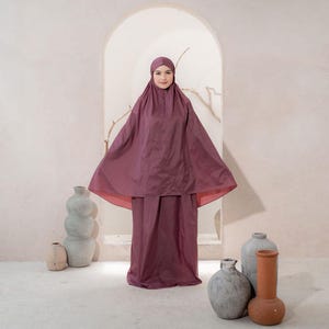 May include: A burgundy-colored prayer garment set, including a head covering and a long skirt. The garment has a flowing design with a wide, cape-like top. The background features neutral-toned vases and an arched doorway.