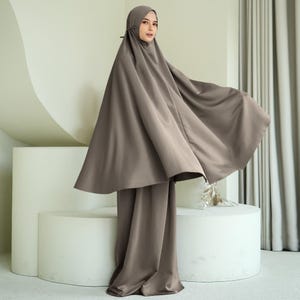 May include: A taupe prayer outfit, comprising a head covering and matching trousers. The outfit has a flowing, modest design. The person is standing in front of a white, curved structure and a neutral-toned background.