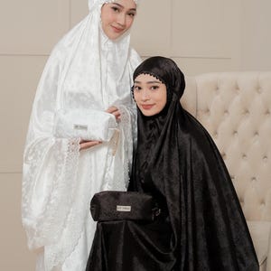 May include: Two women wearing matching prayer garments. One is in white with floral lace trim, holding a matching pouch. The other is in black with a black lace trim. The black garment has the brand name "NIDIA" at the bottom.