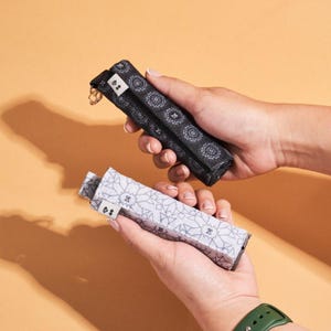 May include: Two patterned cases are held in hands against a peach background. One case is black with a circular design, and the other is white with a geometric pattern. Both cases have a small tag with a heart symbol.