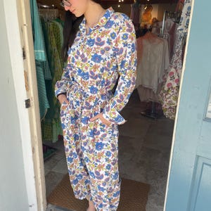 May include: A long-sleeved jumpsuit with a floral pattern in blue, yellow, and pink on a white background. The jumpsuit has a collared neckline, a tie waist, and pockets. The model is wearing silver shoes.