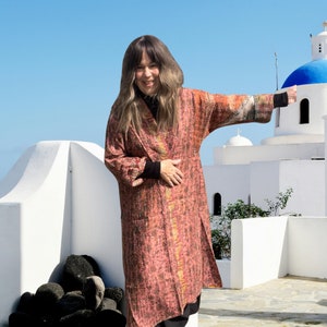 May include: A long, open-front coat in shades of red, brown, and gold. The coat has a textured appearance and a split front. The sleeves have black cuffs. The background features white buildings and a blue sky.