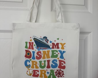 May include: White tote bag with the text "IN MY DISNEY CRUISE ERA" in colorful bubble letters. The design includes a cruise ship, anchor, and ship's wheel. The bag has two white handles.