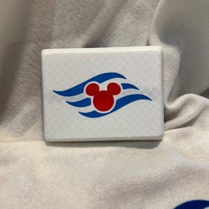May include: A white, rectangular object with a red Mickey Mouse head logo and blue wave accents. The object is set against a white fabric background, showcasing a simple, clean design.