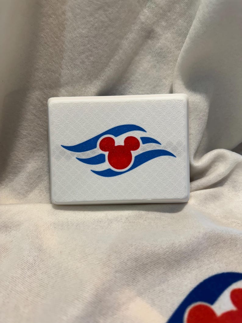 May include: A white, rectangular object with a red Mickey Mouse head logo and blue wave accents. The object is set against a white fabric background, showcasing a simple, clean design.