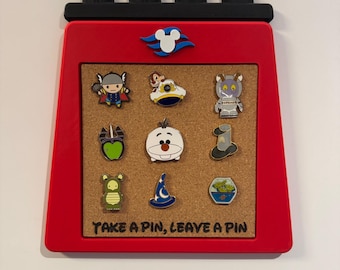 May include: A red, ship-funnel-shaped cork pin board with a black top featuring four smokestacks and a blue and white cruise logo. The board displays nine assorted character pins and the black text, TAKE A PIN, LEAVE A PIN, at the bottom.