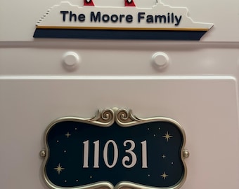 May include: A white door with two decorative signs. The top sign is a ship with the text "The Moore Family". The bottom sign is a dark blue plaque with the number "11031" in white, surrounded by a silver frame.