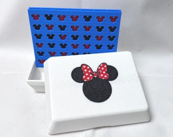 May include: A blue and white game set featuring a Minnie Mouse design. The blue game board has a grid of red and black Mickey Mouse head shapes. The white lid has a black Mickey Mouse head with a red polka dot bow.