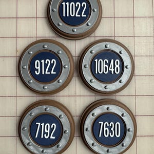 May include: Five circular, brown and silver-toned decorative pieces with white numbers on a navy blue background. The numbers are 11022, 9122, 10648, 7192, and 7630. The pieces have a riveted silver border.