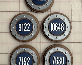 May include: Five circular, brown and silver-toned decorative pieces with white numbers on a navy blue background. The numbers are 11022, 9122, 10648, 7192, and 7630. The pieces have a riveted silver border.