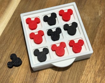 May include: A white square tray with nine Mickey Mouse-shaped game pieces, alternating between red and black. One black piece is separate from the tray. The tray sits on a wooden surface.