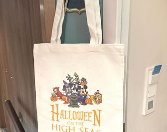 May include: White canvas tote bag featuring a Halloween-themed graphic with cartoon characters and the text "Halloween on the High Seas" in orange. The bag has two white handles and is hanging on a door.