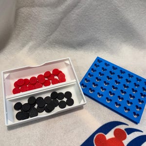May include: A white tray with two compartments contains red and black circular pieces. A blue rectangular piece with Mickey Mouse head cutouts is next to the tray. The items are on a white surface with a Disney Cruise Line logo.