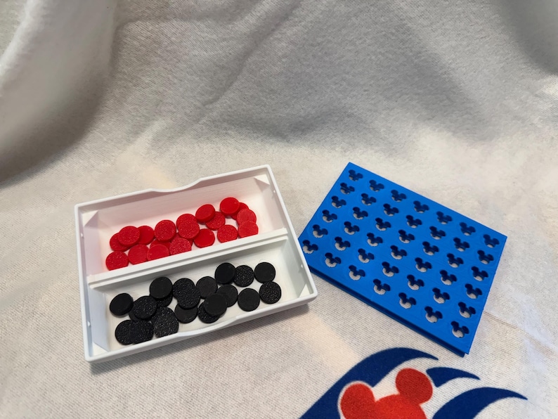 May include: A white tray with two compartments contains red and black circular pieces. A blue rectangular piece with Mickey Mouse head cutouts is next to the tray. The items are on a white surface with a Disney Cruise Line logo.