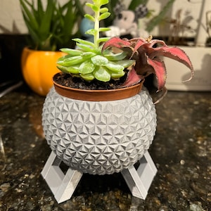 May include: A gray, 3D printed planter shaped like a sphere with a geometric pattern, sitting on a gray, 3D printed stand. The planter holds three succulents, one with green leaves and two with red leaves.