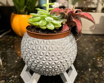 May include: A gray, 3D printed planter shaped like a sphere with a geometric pattern, sitting on a gray, 3D printed stand. The planter holds three succulents, one with green leaves and two with red leaves.