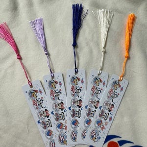 May include: Five white metal bookmarks with colorful tassels. Each bookmark features a cartoon design with Mickey Mouse, Minnie Mouse, and Pluto in nautical outfits. Tassels are pink, purple, blue, white, and orange.