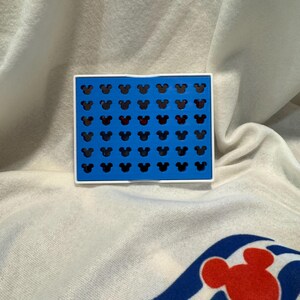 May include: A blue and white rectangular item featuring a grid of black Mickey Mouse head silhouettes, with one row in red. The item rests on a white surface with a Mickey Mouse graphic.