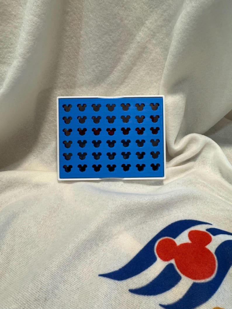 May include: A blue and white rectangular item featuring a grid of black Mickey Mouse head silhouettes, with one row in red. The item rests on a white surface with a Mickey Mouse graphic.