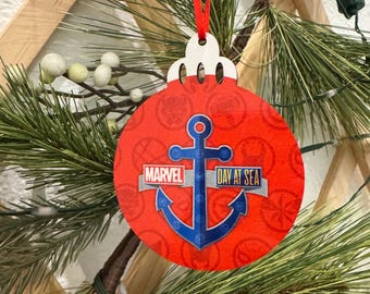 May include: A round, red Christmas ornament with a blue anchor design and the words "Marvel Day at Sea." The ornament is hanging from a red ribbon and is decorated with a nautical theme. The ornament is hanging on a Christmas tree.