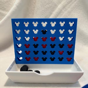 May include: A blue Connect Four game featuring Mickey Mouse head-shaped holes. The game includes white, red, and black counters. The game is set on a white tray.