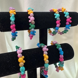 May include: Five beaded bracelets displayed on a black T-bar stand. Each bracelet features a variety of colorful round beads, including solid colors and marbled patterns, interspersed with small three-bead clusters shaped like mouse ears.