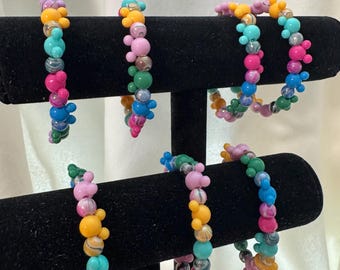 May include: Five beaded bracelets displayed on a black T-bar stand. Each bracelet features a variety of colorful round beads, including solid colors and marbled patterns, interspersed with small three-bead clusters shaped like mouse ears.