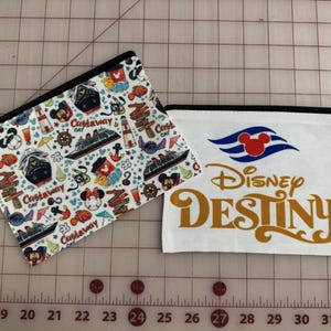 May include: Two zippered pouches. One features a white background with a pattern of Mickey Mouse, cruise ships, and the text "Castaway Cay." The other is white with the Disney Cruise Line logo and "Disney Destiny" in gold.
