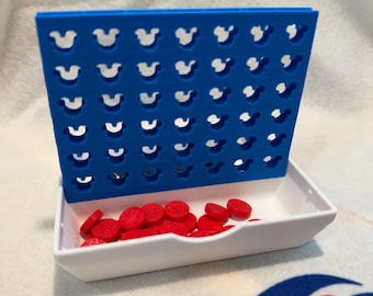 May include: A blue Connect Four game board with Mickey Mouse head-shaped cutouts. The game includes a white tray filled with red game pieces. The game is set on a white surface with a red and blue design.