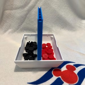 May include: A white plastic tray with a blue divider, containing black and red circular game pieces. A blue vertical piece is in the centre. The tray rests on a white cloth with a red and blue Mickey Mouse design.