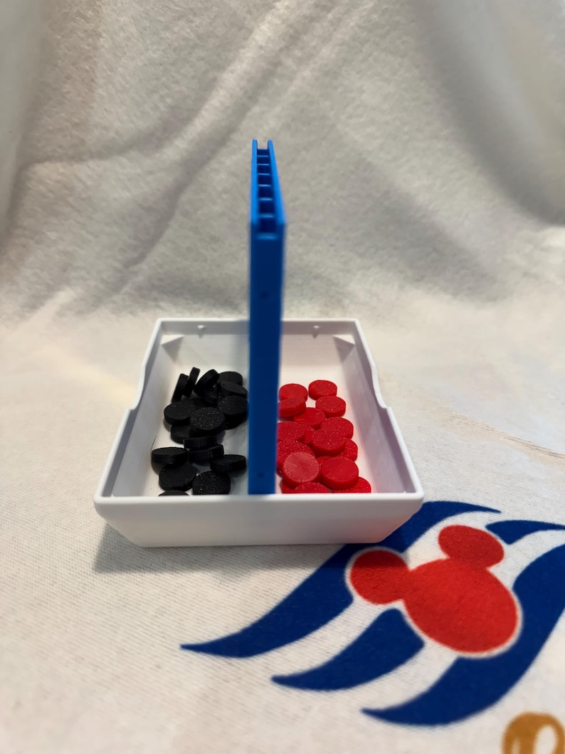 May include: A white plastic tray with a blue divider, containing black and red circular game pieces. A blue vertical piece is in the centre. The tray rests on a white cloth with a red and blue Mickey Mouse design.
