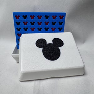 May include: A white, square-shaped game box with a black Mickey Mouse silhouette on the lid. A blue game board with Mickey Mouse head cutouts is behind the box. The game board has a red interior.