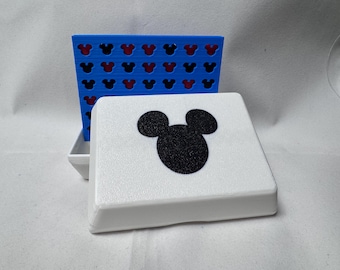 May include: A white, square-shaped game box with a black Mickey Mouse silhouette on the lid. A blue game board with Mickey Mouse head cutouts is behind the box. The game board has a red interior.