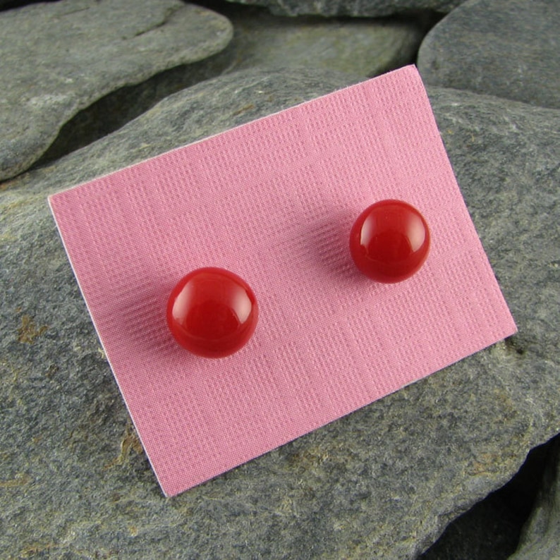 May include: A pair of bright red, round stud earrings. The earrings are displayed on a textured pink card, set against a backdrop of gray stones. The earrings have a glossy finish.