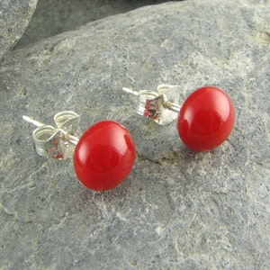 May include: A pair of red, oval-shaped stud earrings with silver posts and backs. The earrings are set against a gray stone surface, showcasing their vibrant color and simple design. The earrings are small and appear to be lightweight.