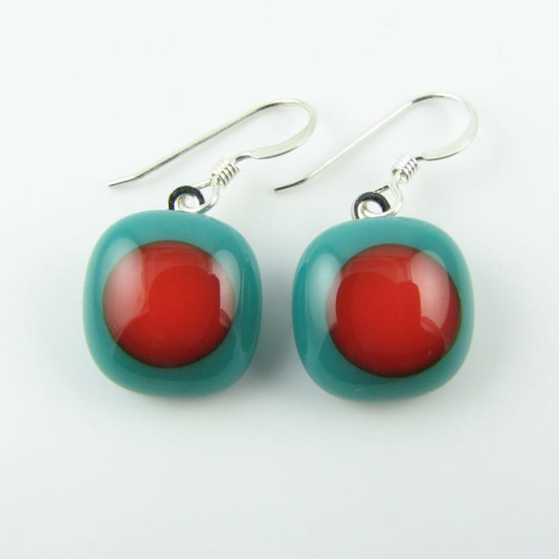 May include: Teal blue glass earrings with a red circle in the center. The earrings have silver hooks.