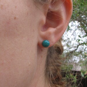 May include: A close-up of a teal-colored stud earring. The earring is a small, round sphere, set against the backdrop of a person's ear and skin. The earring's color is a vibrant teal, providing a pop of color.