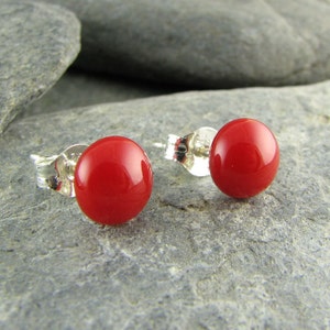 May include: A pair of red, round stud earrings. The earrings have a glossy finish and are set on silver posts. The earrings are displayed on a gray, textured surface.