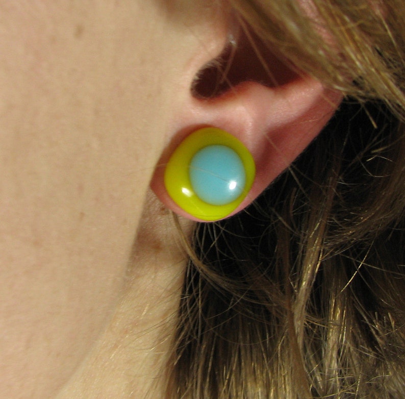 May include: A yellow and blue glass earring with a round shape. The earring has a yellow outer ring and a blue inner circle.