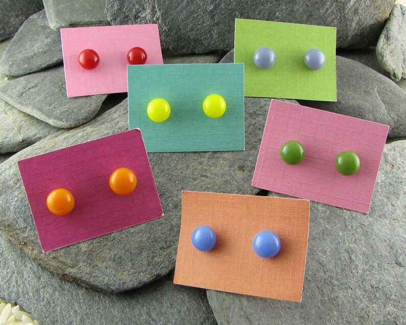 May include: A collection of colorful stud earrings displayed on small, textured cards. The earrings are in various colors, including red, yellow, green, orange, and blue. The cards are pink, green, purple, and orange. The earrings are displayed on a gray stone surface.