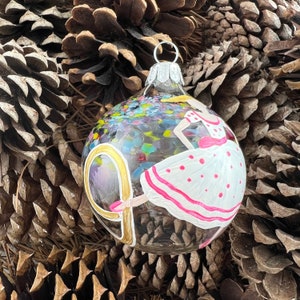 Könnte beinhalten: Transparenter Weihnachtsbaumschmuck aus Glas mit einer gemalten Ballerina in einem weißen Kleid mit rosa Punkten und Verzierungen. Der Schmuck hat eine silberne Metallkappe und einen bunten, gesprenkelten Hintergrund. Die Ballerina hält eine goldfarbene Requisite.