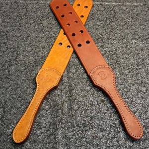 May include: Two leather paddles in shades of brown and tan are displayed against a textured grey background. The paddles have a handle and a rectangular striking surface with several holes. The handles have a decorative embossed design.