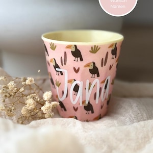 Könnte beinhalten: Ein rosa Becher mit einem wiederkehrenden Muster aus Tukanen, kleinen braunen Herzen und grünem Blattwerk. Der Name Jana ist in großen weißen Schreibschriftbuchstaben auf der Vorderseite aufgedruckt. Ein runder Aufkleber oben rechts enthält den Text Personalisiert mit deinem Wunsch Namen.
