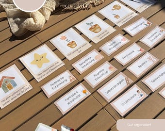 May include: A collection of rectangular cards featuring German text and illustrations, including a star, a house, and potted plants. The cards are arranged on a wooden surface alongside a white fabric cloth, a glass bottle, and a chocolate bar.
