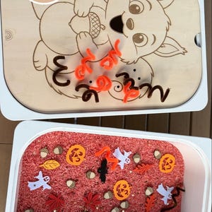 Könnte beinhalten: Ein Sensorik-Set mit einem hellen Holzdeckel, der ein lasergraviertes Eichhörnchen mit Eichel und den Text Perfekte Beschäftigung zeigt. Der untere Behälter ist mit rotem Reis, orangefarbenen Pfeifenreinigern und Filzfiguren wie Kürbissen, Geistern, Fledermäusen, Blättern und Eicheln gefüllt.