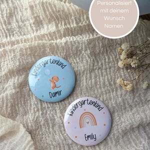 Könnte beinhalten: Zwei runde Ansteckbuttons mit glänzender Oberfläche. Ein hellblauer Button zeigt eine Tiger-Illustration, einen Luftballon und den Text Kindergartenkind Damir. Der andere hellviolette Button zeigt eine Regenbogen-Grafik und den Text Kindergartenkind Emily.