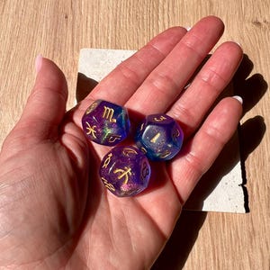 Op de afbeelding: Drie iriserende, veelzijdige dobbelstenen met gouden dierenriemtekens worden in een hand gehouden. De dobbelstenen zijn paars en blauw, met een glinsterend, glinsterend effect. De dobbelstenen liggen op een kleine, vierkante, witte tegel.